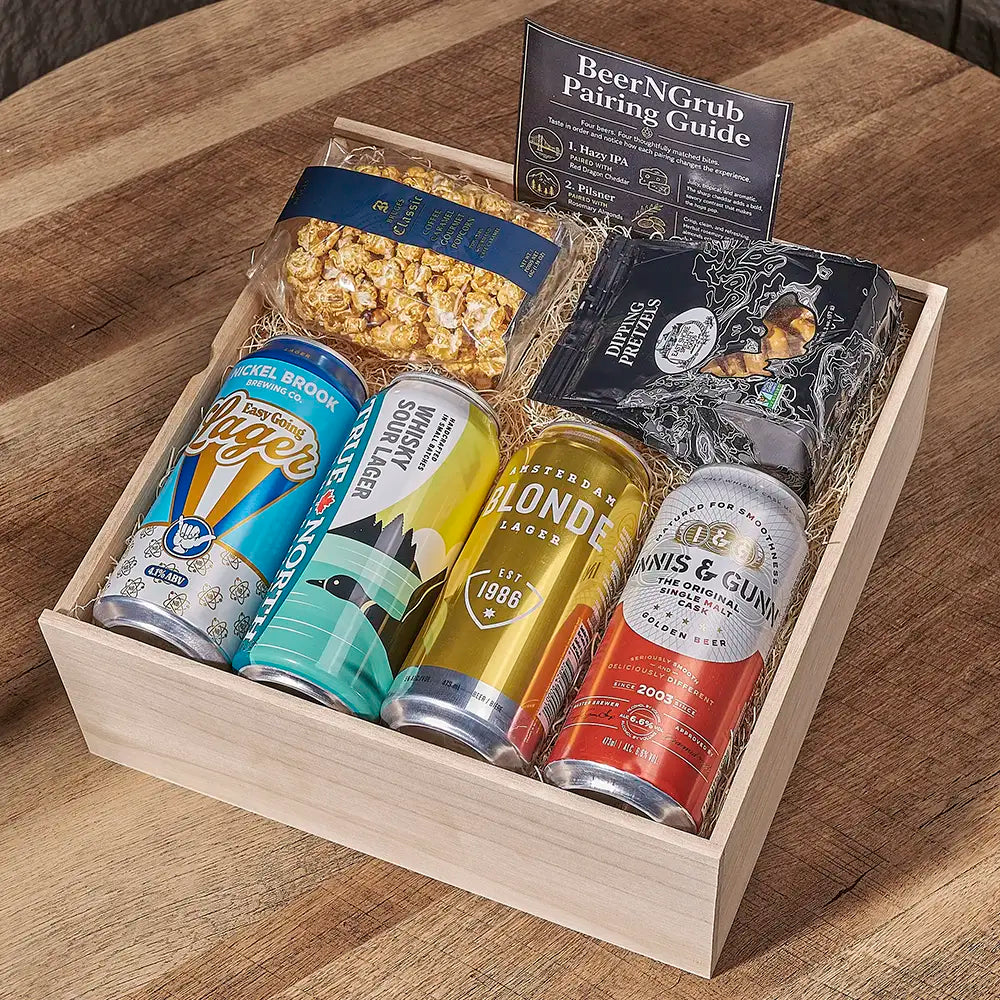 A wooden gift box filled with assorted beer pairings, including four canned beers, a bag of popcorn, a black packaged snack, and a booklet titled BeerNGrub Pairing Guide, arranged on a wooden table.