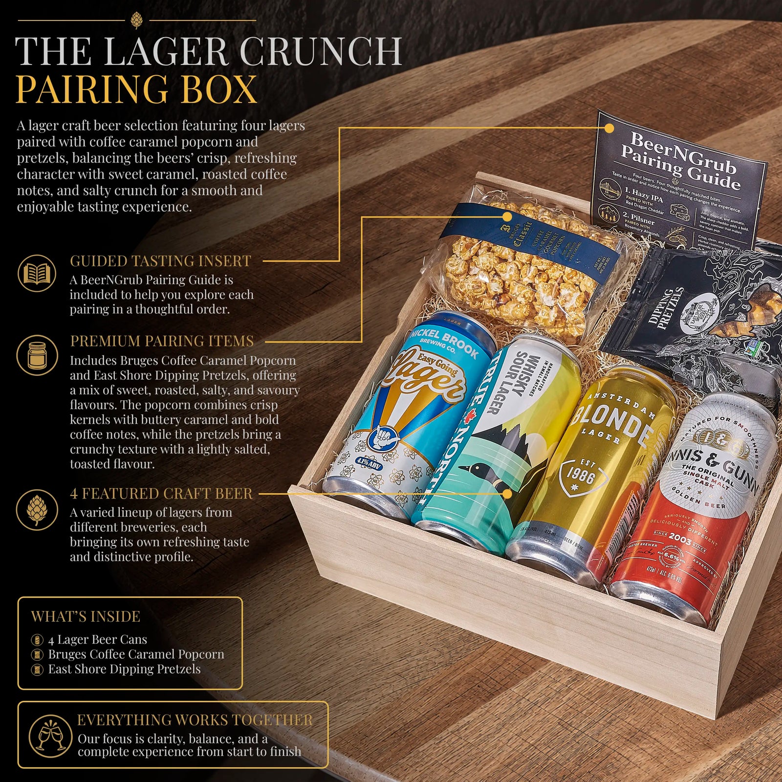 A curated beer pairing box labeled 'The Lager Crunch Pairing Box' displayed in a wooden tray on a wooden tabletop. The box includes multiple beer cans, a bag of caramel popcorn, pretzels, and a Beer or Grub Pairing Guide booklet.