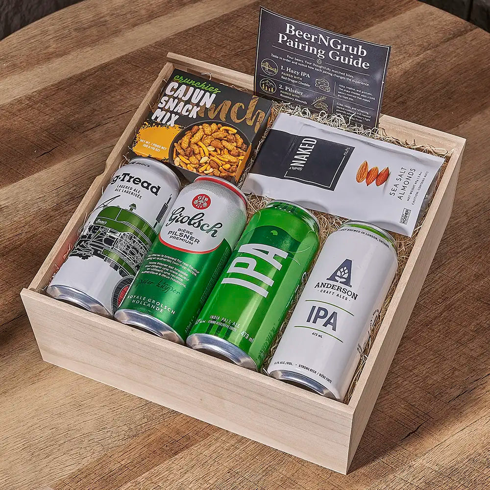 A wooden gift box filled with beer cans, a bag of Cann Smack Mix, a packaged snack, and a BeerNGrub pairing guide, arranged on a wooden table.