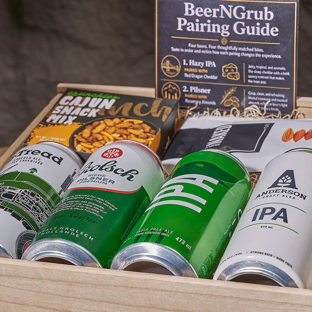 A curated beer and snack gift box arranged in a wooden tray, featuring canned beers from Porter, KBS, and Anderson Craft Ale, a bag of Cajun Snack Mix, and a BeerNGrub pairing guide card standing behind the items.