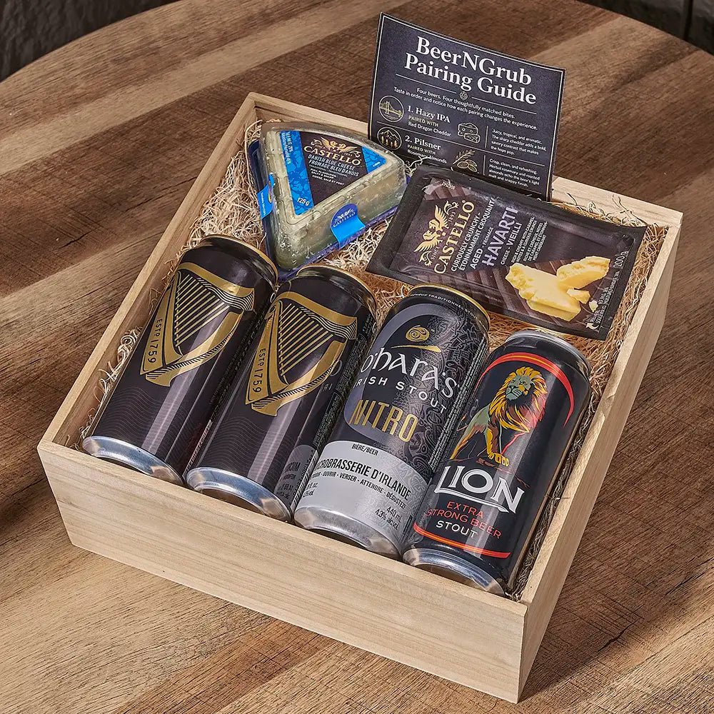 A wooden gift box filled with beer and food items, including two stout cans, a can of Nitro beer, a packaged cheese wedge, a cheese stick, and a pairing guide booklet, arranged on a wooden table.