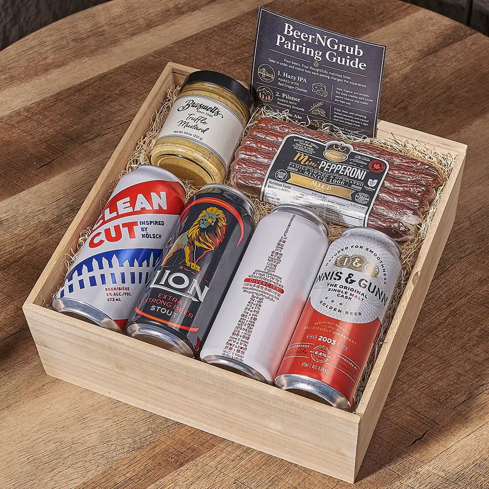A wooden gift box filled with assorted food items and a beer pairing guide, placed on a wooden table. The box contains a jar of honey mustard, two canned beers, a pack of pepperoni, and a bottle of wine.