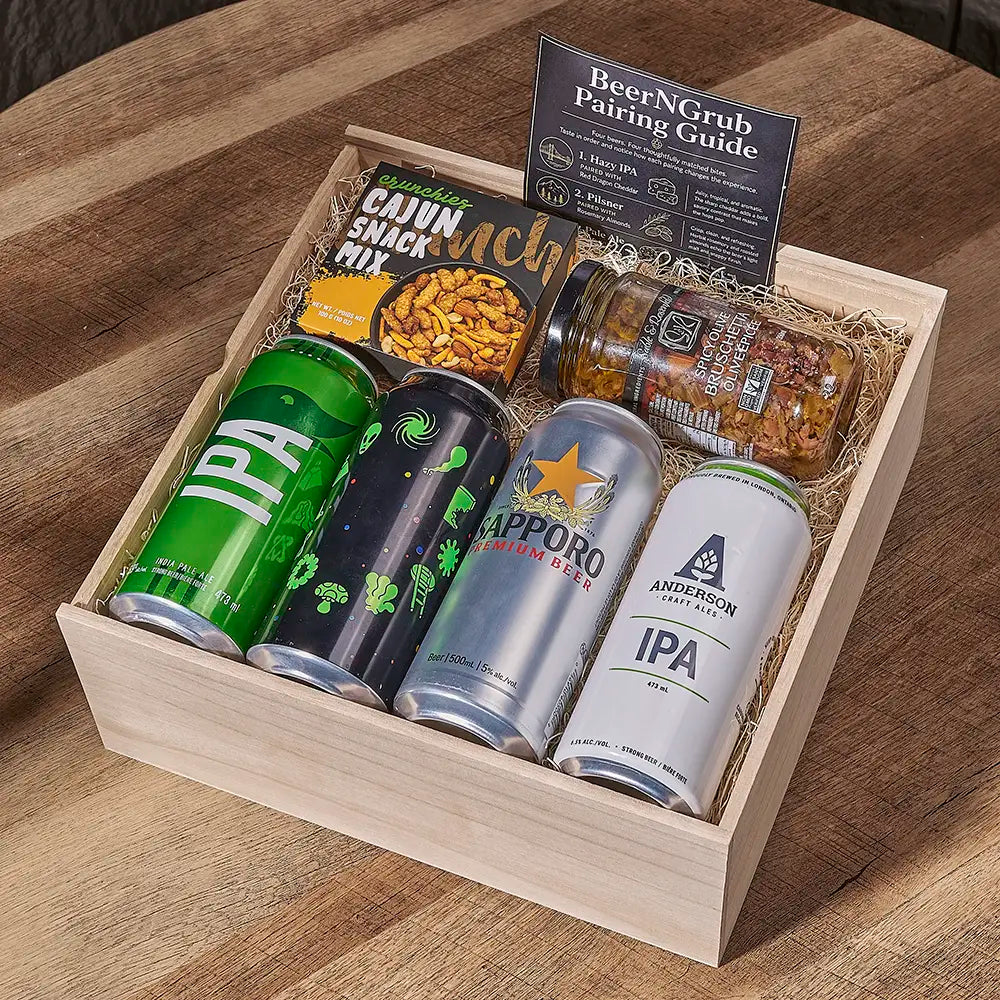 A wooden gift box filled with assorted beer and snack items on a wooden table. The set includes four canned beers from Anchor Brewing Company, Capporo, and Sierra Nevada, a jar of chutney, a bag of Cajun Snack Mix, and a pairing guide card.