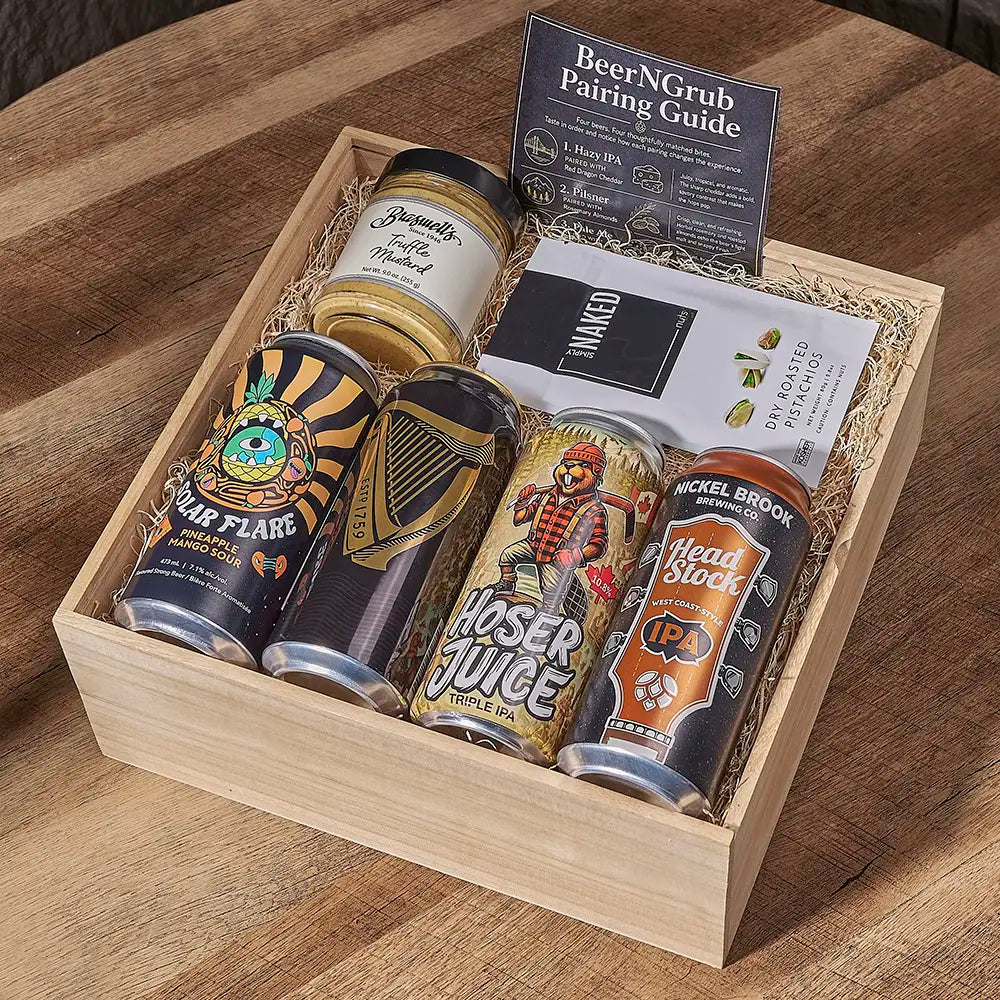 A wooden gift box filled with assorted beer cans, a jar, a pairing guide, and a card, arranged on a wooden table. Visible brands on the products include Hoser Juice, Buckel Brook, and Headstock.