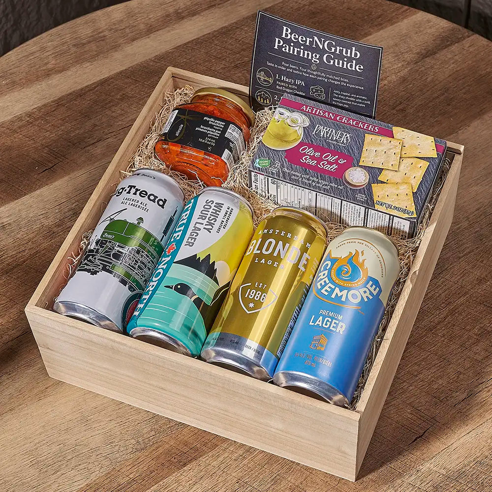 A wooden gift box filled with assorted food and drinks on a wooden table. The box contains four canned beers, a jar of pickled vegetables, a box of crackers, and a booklet titled BeerNGrub Pairing Guide.