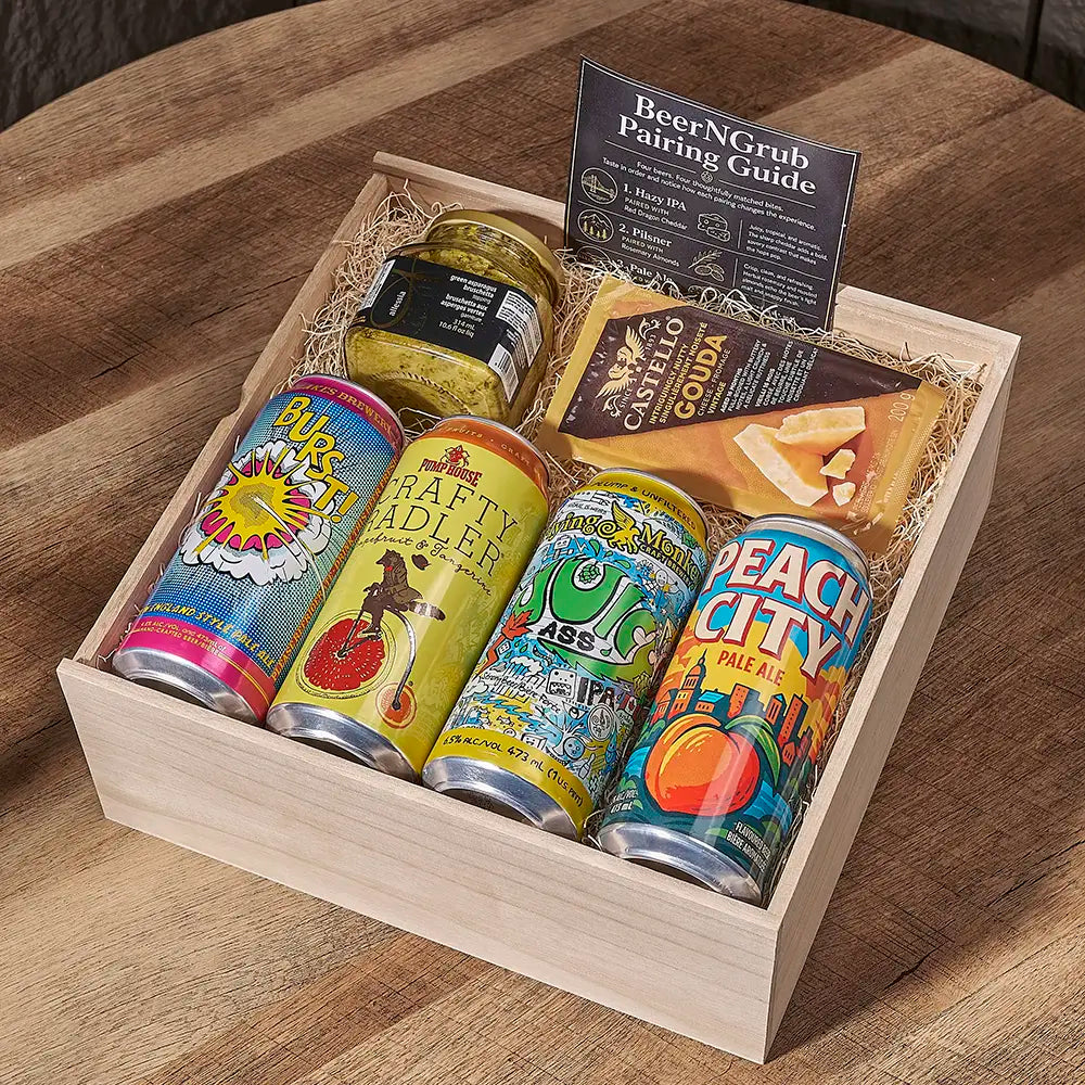 A wooden gift box filled with assorted snacks and drinks on a round wooden table. The box contains four colorful canned beverages, a jar of pickled vegetables, a packaged cheese wedge labeled Cash's Couza, and a black card titled BeerNCrub Pairing Guide.