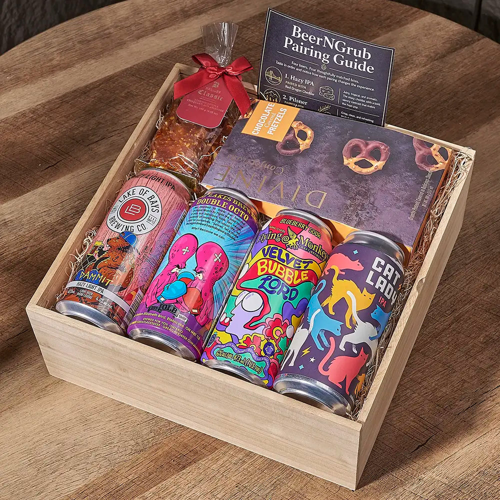 A wooden gift box filled with assorted snacks and drinks on a wooden table. The box contains colorful canned beverages, pretzel snacks, a bag of chips wrapped in a red ribbon, and a card titled 'BeerNGrub Pairing Guide'.
