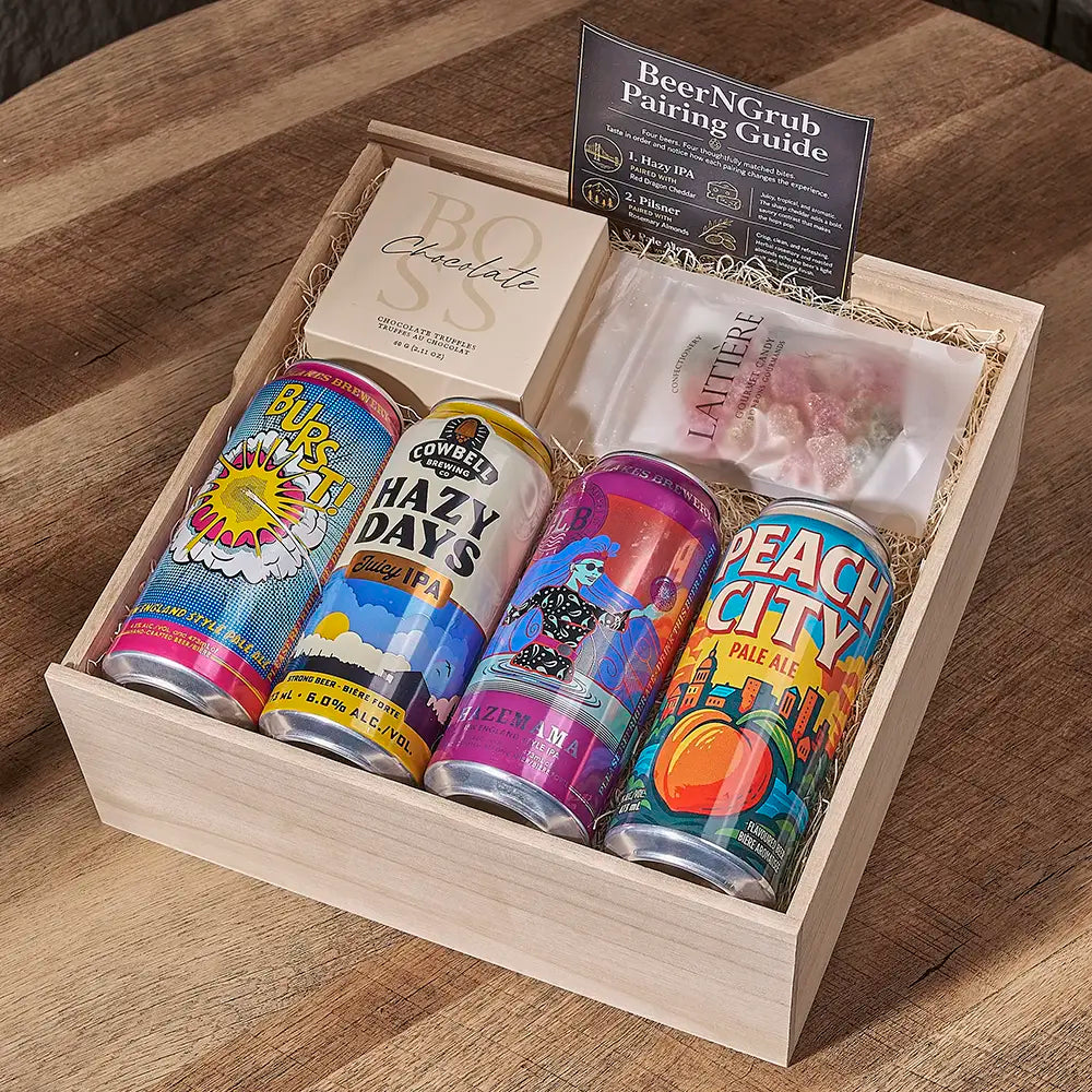 A wooden gift box filled with beer cans, a chocolate box, a plastic bag of snacks, and a booklet titled 'BeerNGrub Pairing Guide' on a wooden table.