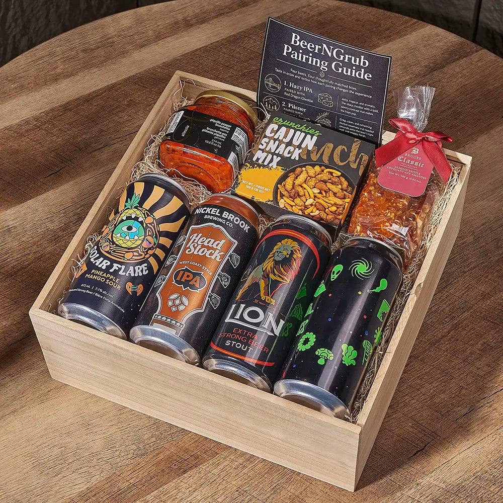A wooden gift box filled with assorted food and drinks, including four craft beer cans, a jar of orange-red sauce, a bag of seasoning snack mix, and a clear bag of marinated onions, arranged on a wooden table. Visible brands include Beel Brook, Lion, Headstock, and Star Flare.