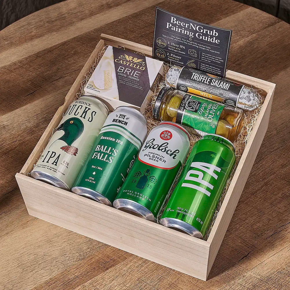 A wooden gift box filled with beer cans, a brie cheese wedge, a truffle salami stick, and a pairing guide, photographed on a wooden table.
