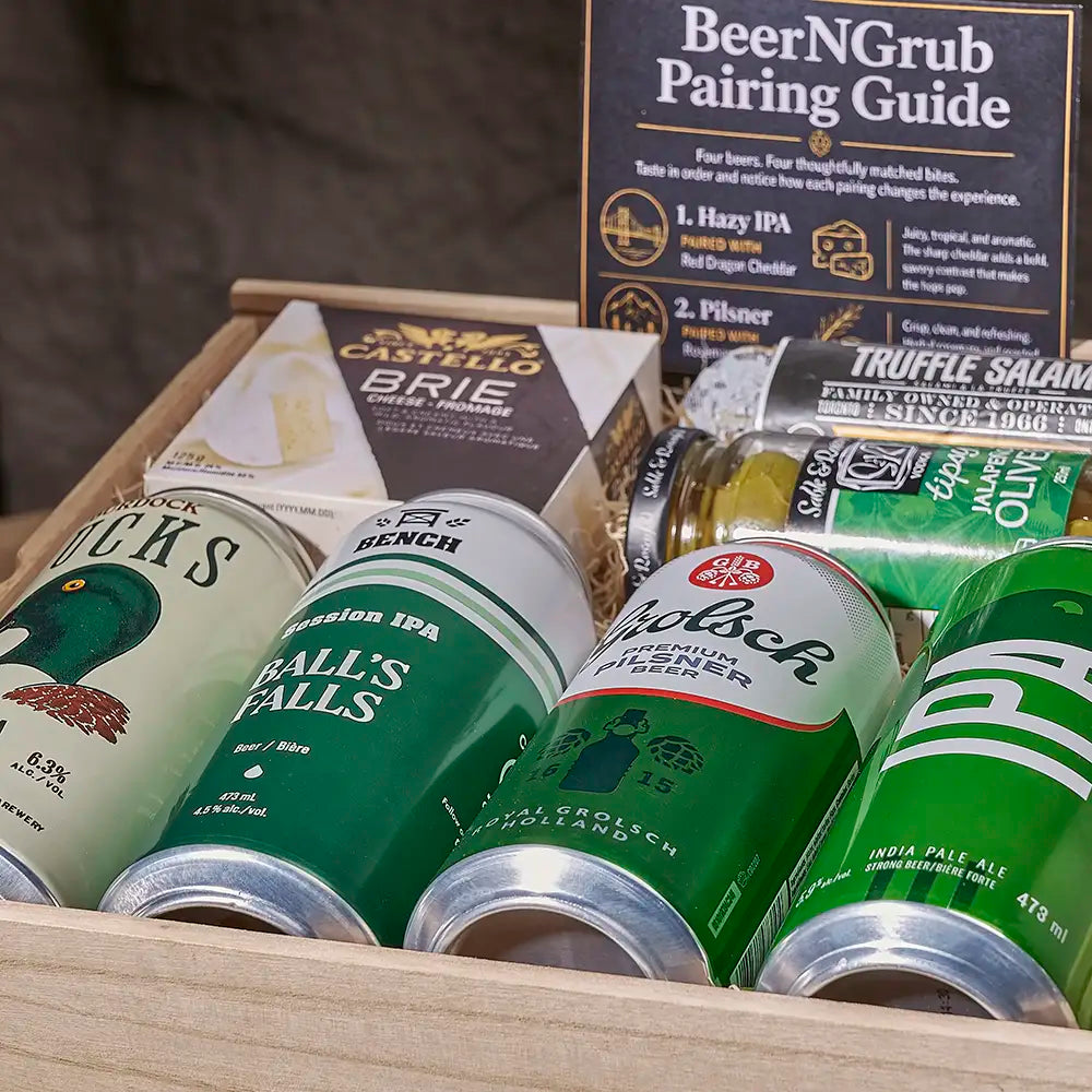 A wooden gift box filled with beer cans and a printed pairing guide titled 'BeerNGrub Pairing Guide.' Visible products include green and white canned beers such as Ball's Falls, Michel, and Truffe Salame, plus a Castello Brie box.