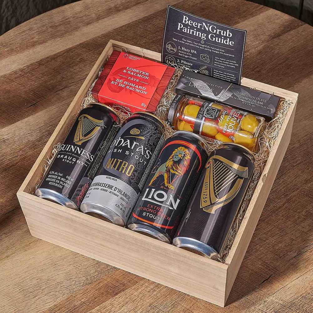 A wooden gift box filled with assorted beer cans, a red packet, and a black pairing guide, photographed on a wooden table. Visible brands on the cans include Guinness, D'Ata's, Lion, and Sam Adams.